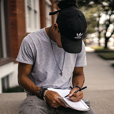 person in black adidas cap sitting on bench writing on notebook